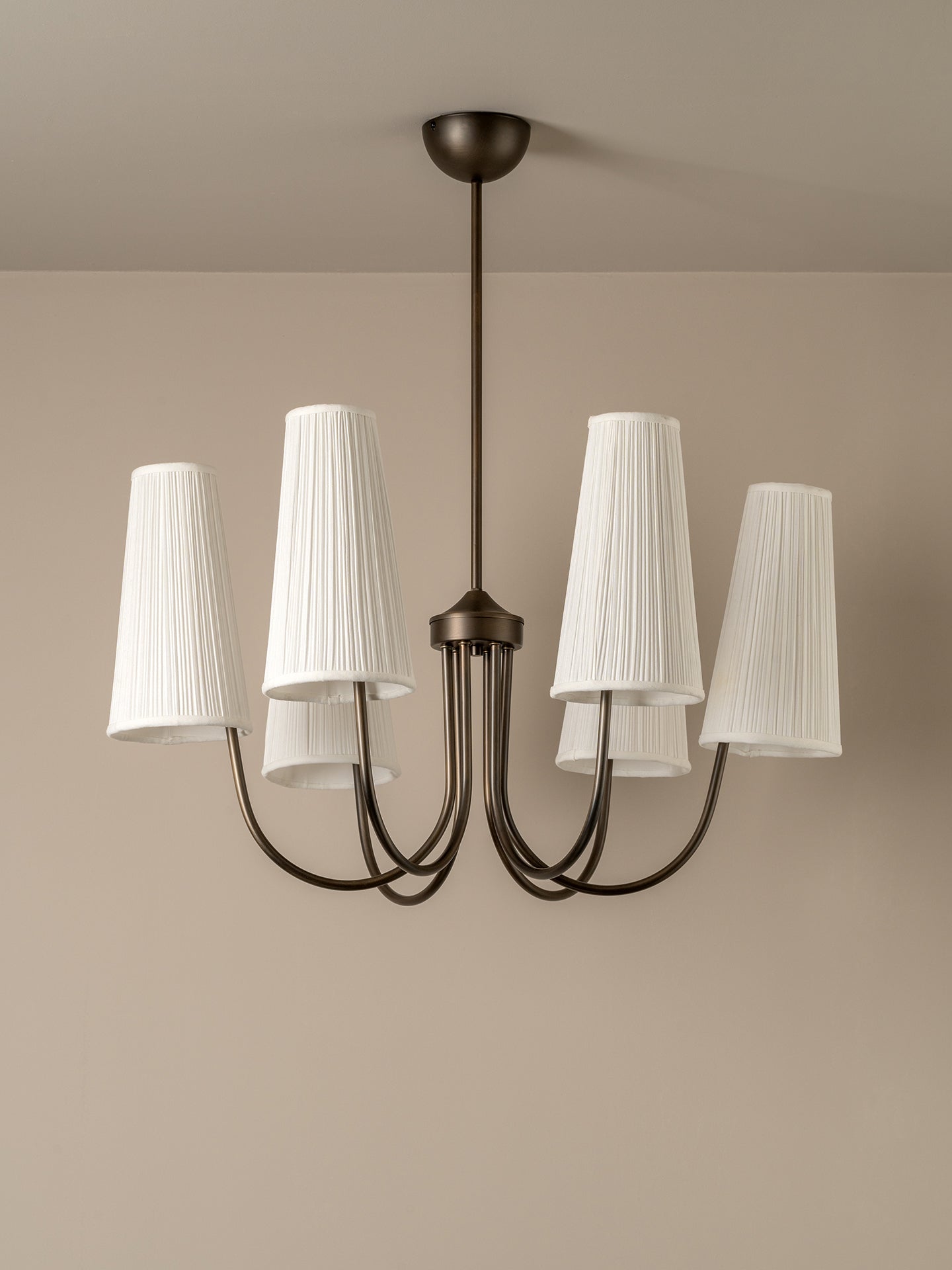 A modern five-light chandelier with a dark bronze metal frame and white pleated drum shades, shown hanging against a neutral beige wall and ceiling.