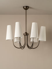 A modern five-light chandelier with a dark bronze metal frame and white pleated drum shades, shown hanging against a neutral beige wall and ceiling.