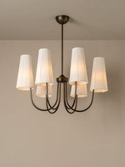 A six-light chandelier with a dark bronze metal frame and white pleated fabric shades, shown illuminated against a neutral beige wall and ceiling.