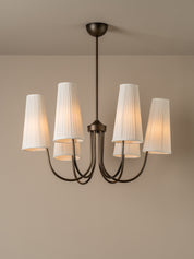 A five-light chandelier with a dark bronze metal frame and white pleated fabric shades, shown illuminated against a neutral beige wall and ceiling.