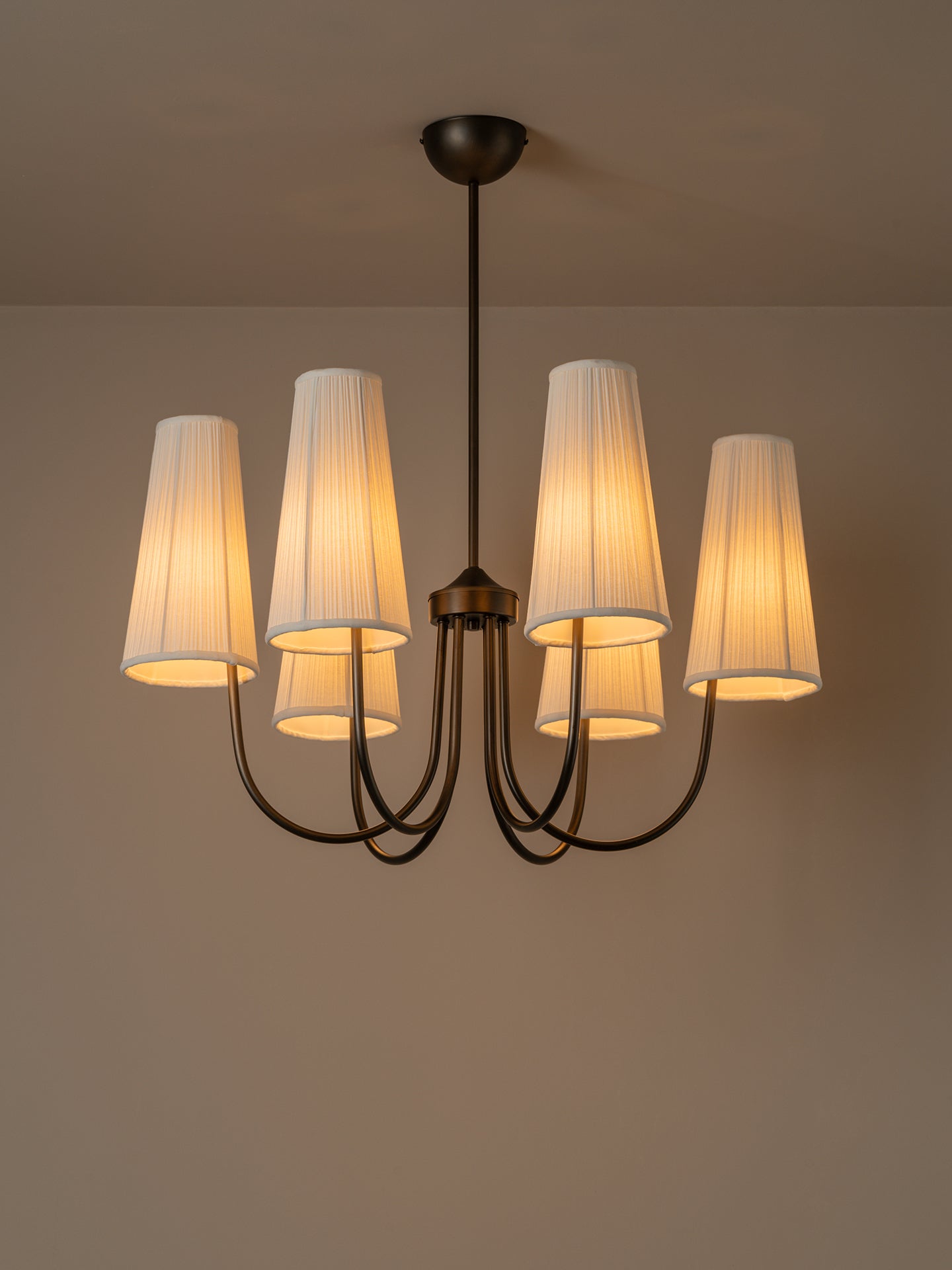 A five-light chandelier with a dark metal frame and pleated off-white fabric shades, glowing with a warm yellow light against a neutral beige wall and ceiling.