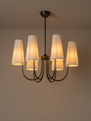 A five-light chandelier with a dark metal frame and pleated off-white fabric shades, glowing with a warm yellow light against a neutral beige wall and ceiling.