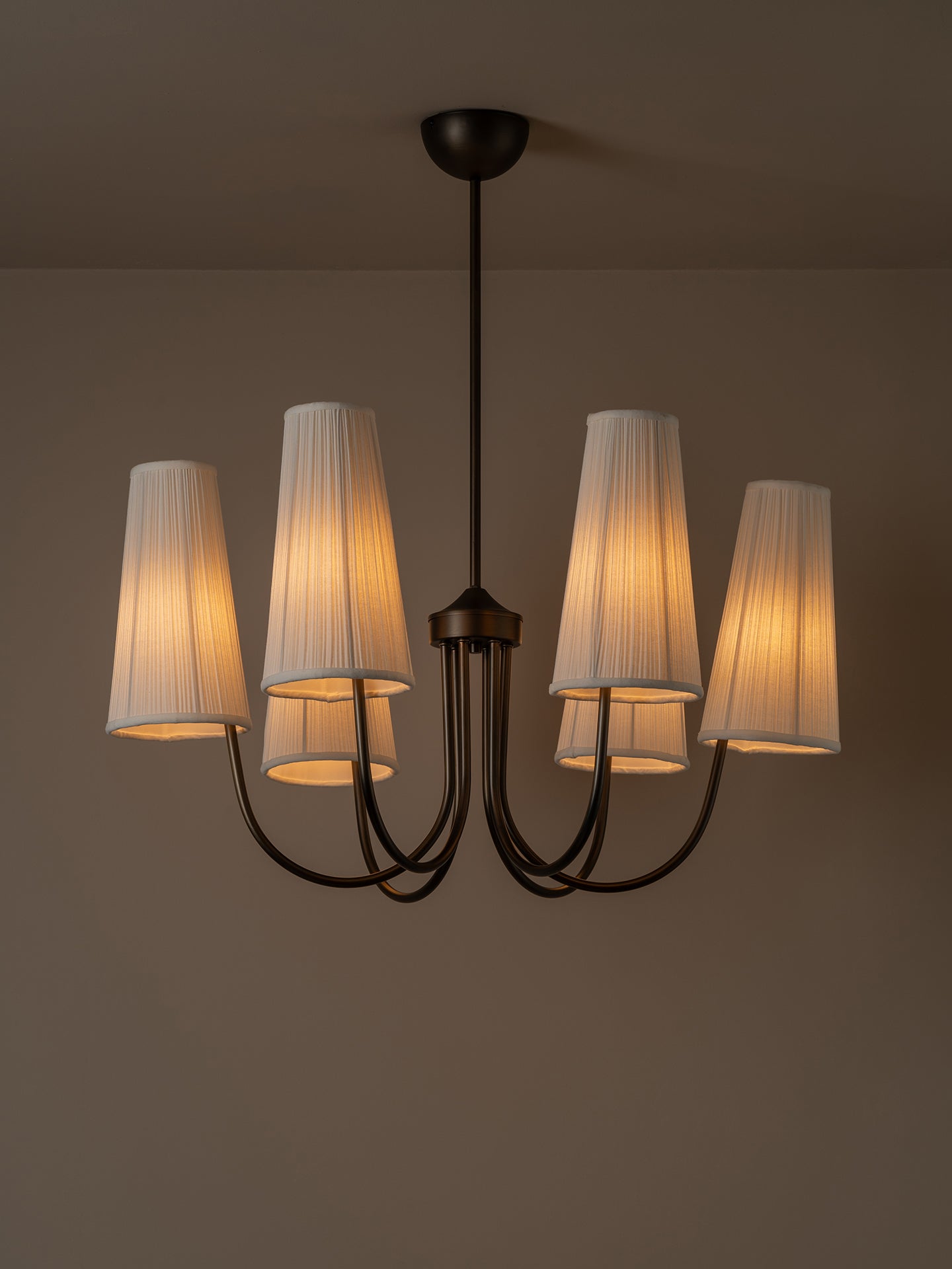 A six-light chandelier with a dark metal frame and pleated off-white fabric shades, illuminated with a warm glow against a neutral brown wall.