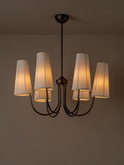 A six-light chandelier with a dark metal frame and pleated off-white fabric shades, illuminated with a warm glow against a neutral brown wall.