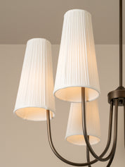 A close-up of a multi-light chandelier with three tapered white pleated fabric shades and a dark bronze metal frame, shown against a neutral beige wall and ceiling.