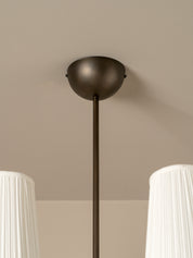A close-up of a modern ceiling light fixture with a matte dark bronze dome canopy and a matching vertical rod, shown installed on a beige ceiling with white pleated lampshades partially visible at the bottom corners.