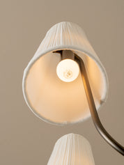 A close-up of a modern floor lamp with a pleated white fabric shade and a curved dark metal arm, shown lit against a warm beige background.