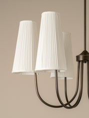 A close-up of a multi-light chandelier with three white pleated drum shades and curved dark metal arms against a warm beige background.