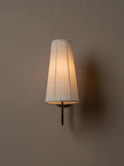 A modern wall-mounted sconce with a pleated white fabric shade, illuminated with a warm glow against a solid taupe wall.