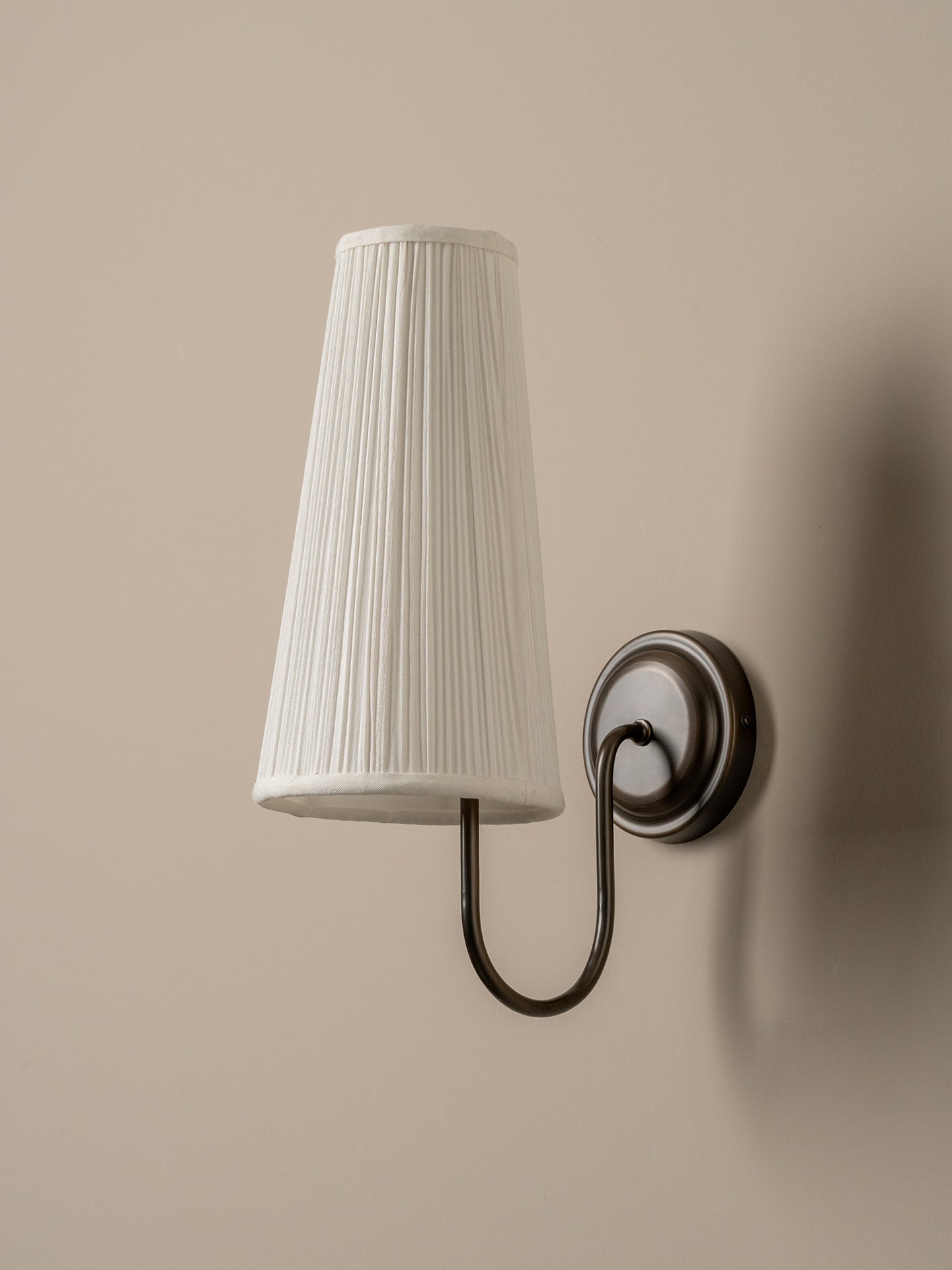 A wall-mounted sconce with a tapered white pleated fabric shade and a dark metal curved arm, shown against a warm beige wall.