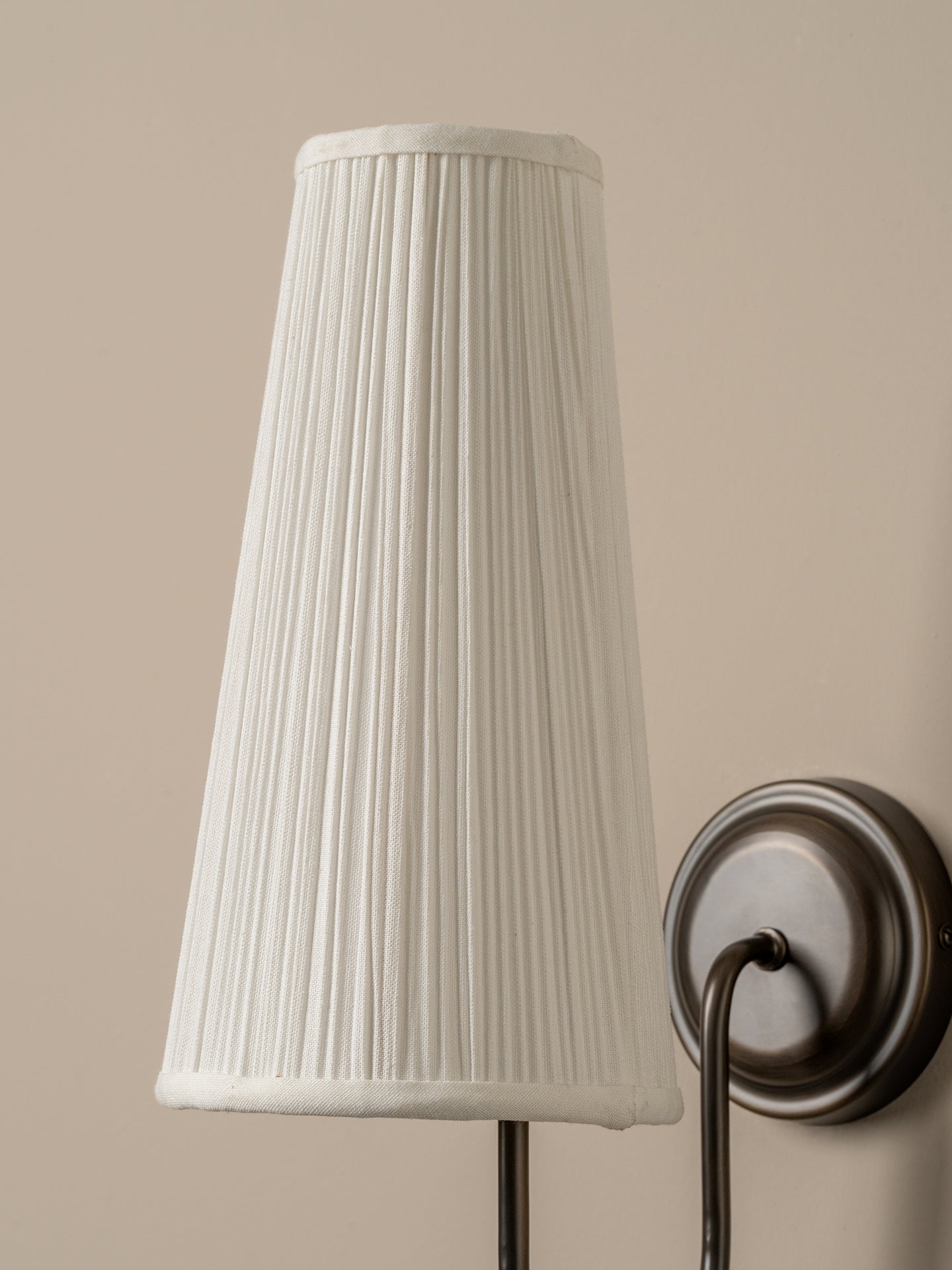 A pleated fabric lampshade in off-white, shown hanging from a dark metal wall sconce against a warm beige wall.