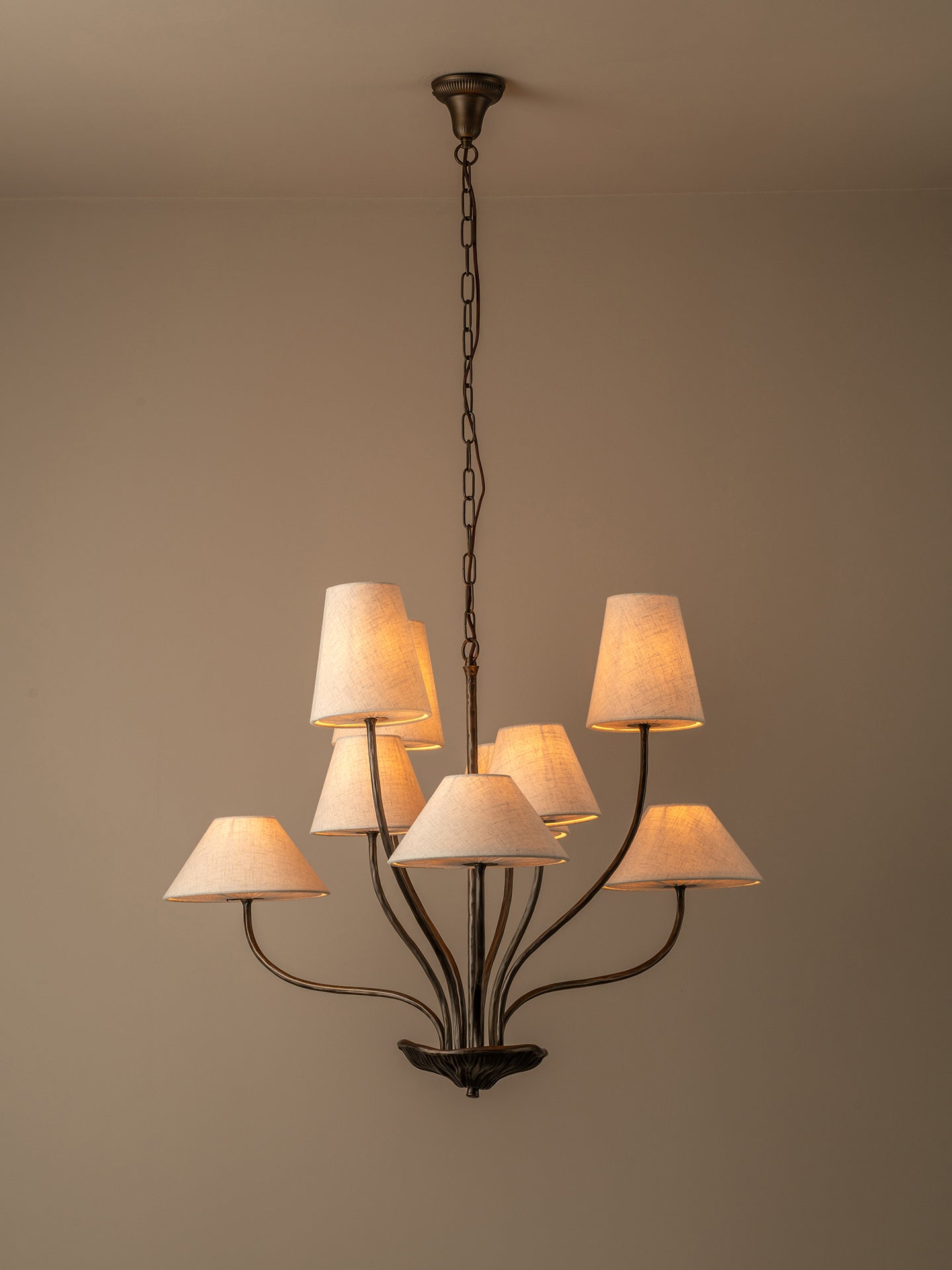 A multi-arm chandelier with a dark metal frame and eight warm white fabric drum shades, hanging from a ceiling against a plain beige wall.