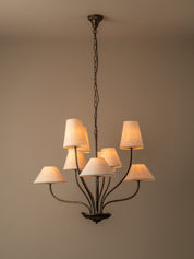 A multi-arm chandelier with a dark metal frame and eight warm white fabric drum shades, hanging from a ceiling against a plain beige wall.