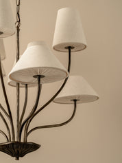 A close-up of a multi-arm chandelier with textured off-white fabric drum shades and a dark metal frame, set against a solid beige background.