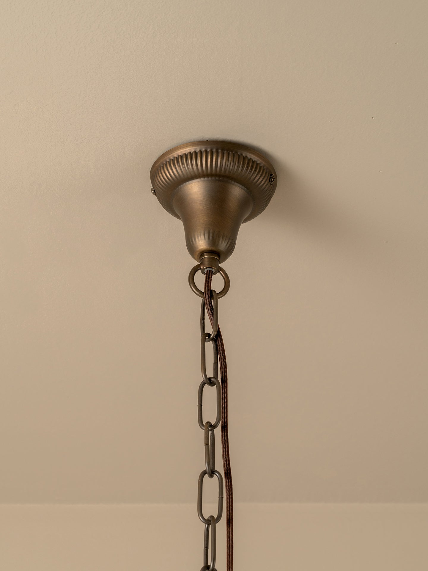 An antique bronze ceiling light fixture canopy with a decorative ribbed top and a hanging chain, shown mounted on a beige wall.