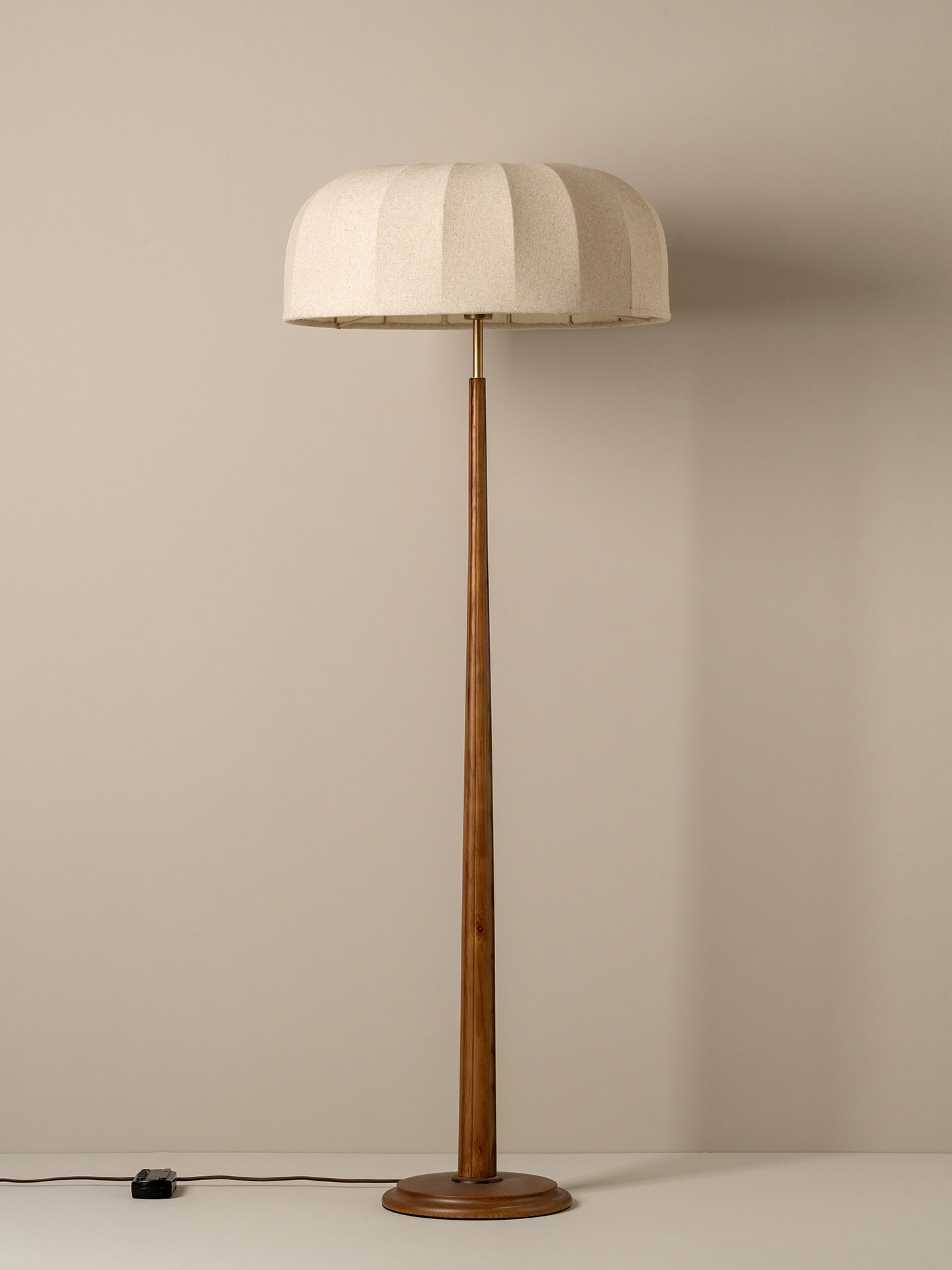 A vintage-style floor lamp with a pleated off-white fabric drum shade and a slender brown wooden pole and base, set against a plain beige wall.