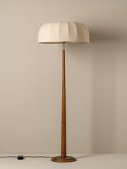 A vintage-style floor lamp with a pleated off-white fabric drum shade and a slender brown wooden pole and base, set against a plain beige wall.