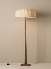 A modern floor lamp with a pleated beige fabric drum shade, a slender dark brown wooden stem, and a round wooden base, set against a neutral beige wall.