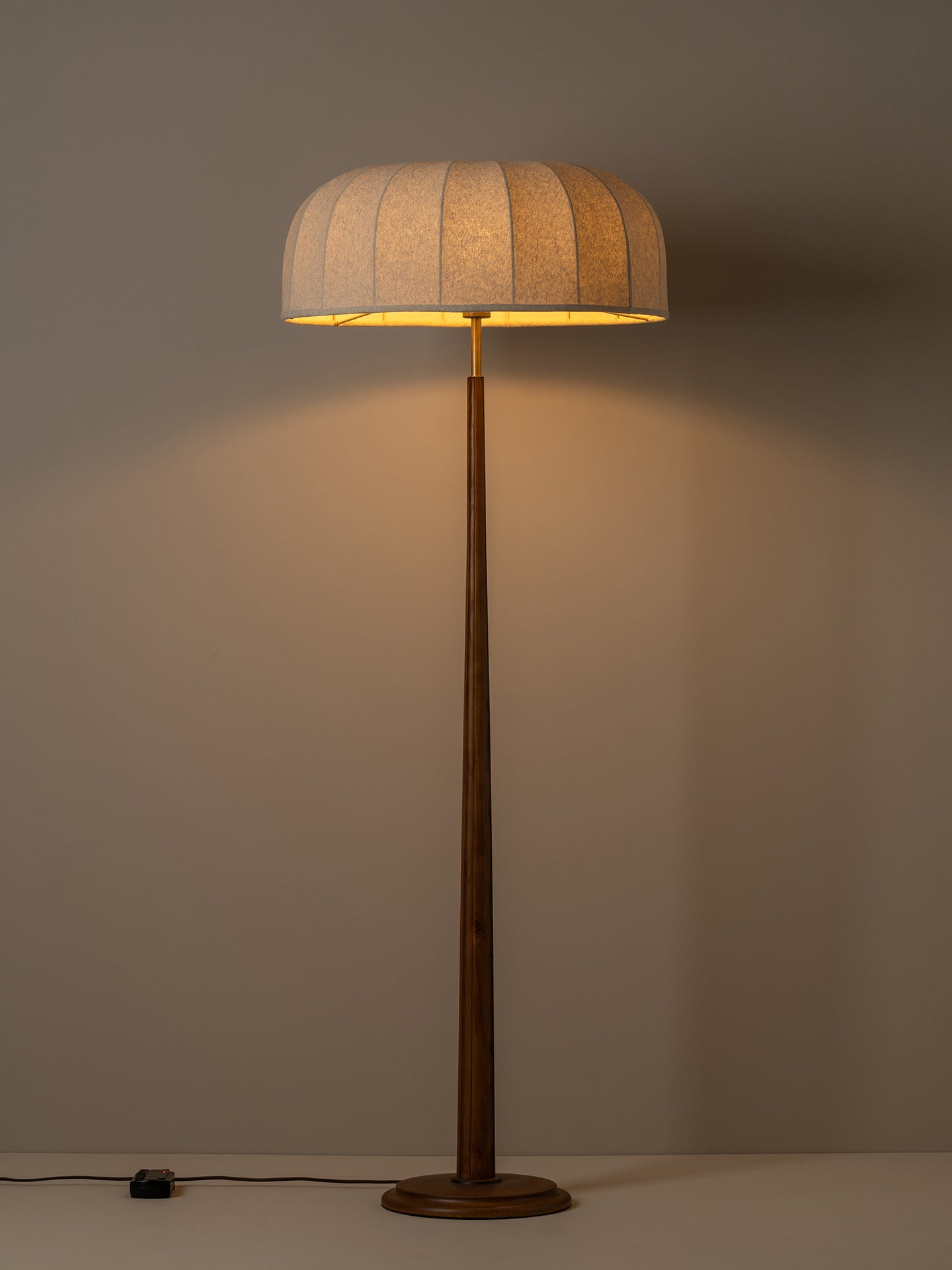 A modern floor lamp with a pleated beige fabric drum shade and a dark brown wooden pole and base, illuminated against a warm neutral background.