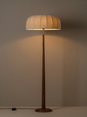 A modern floor lamp with a pleated beige fabric drum shade and a dark brown wooden pole and base, illuminated against a warm neutral background.