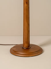 Close-up of a wooden floor lamp base and central pole, showing a warm medium-brown wood grain finish against a plain beige background.