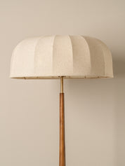 A modern floor lamp with a large, pleated dome-shaped shade in a textured cream fabric, supported by a slim wooden pole and a brass finial, set against a solid beige background.