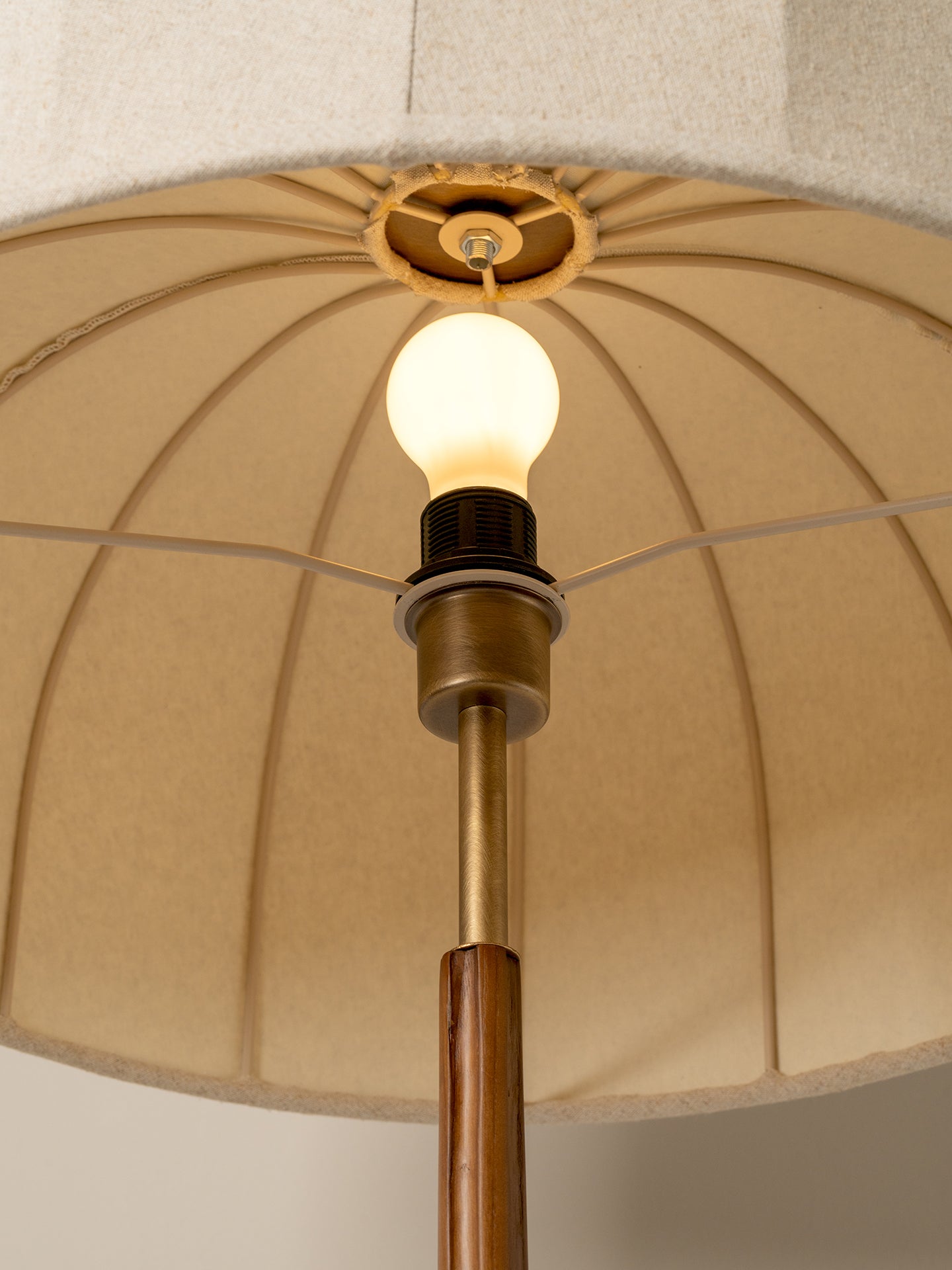 Close-up of a floor lamp with a large beige pleated fabric drum shade, a brass-toned metal stem, a warm glowing light bulb, and a dark collar.