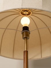 Close-up of a floor lamp with a large beige pleated fabric drum shade, a brass-toned metal stem, a warm glowing light bulb, and a dark collar.