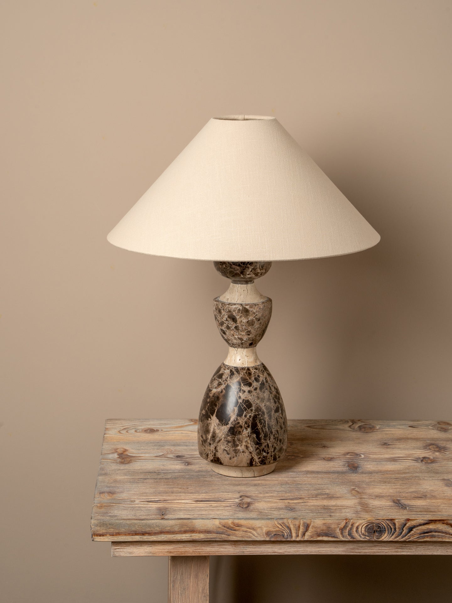 A table lamp with a cream-colored fabric cone shade and a sculptural, bulbous base made of polished stone in earthy brown, beige, and black marbled tones, displayed on a rustic wooden table against a solid beige wall.