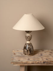 A table lamp with a cream-colored fabric cone shade and a sculptural, bulbous base made of polished stone in earthy brown, beige, and black marbled tones, displayed on a rustic wooden table against a solid beige wall.