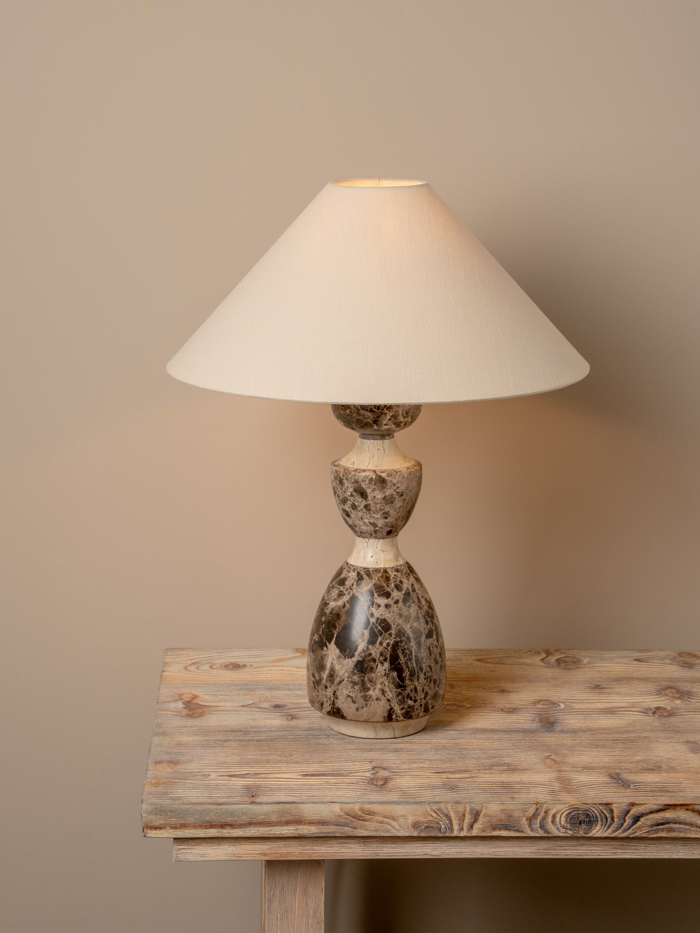 A table lamp with a cream-colored fabric drum shade and a sculptural, bulbous base made of polished brown and beige marbled stone, shown resting on a rustic wooden table against a solid beige wall.