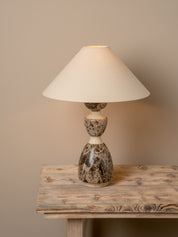 A table lamp with a cream-colored fabric drum shade and a sculptural, bulbous base made of polished brown and beige marbled stone, shown resting on a rustic wooden table against a solid beige wall.