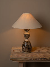 A table lamp with a warm white fabric drum shade and a dark brown and beige marbled stone base, shown lit on a rustic wooden table against a neutral brown wall.