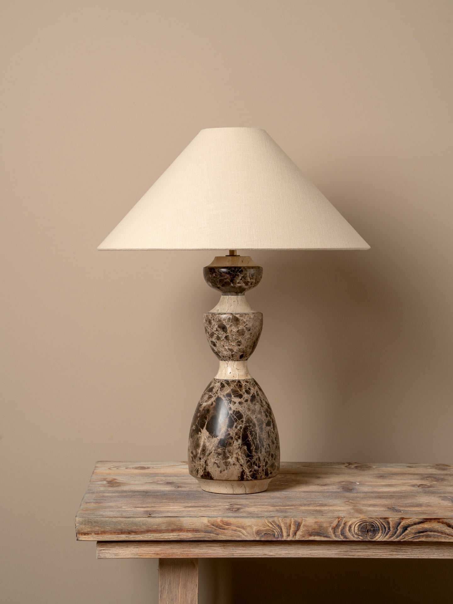 A table lamp with a cream fabric drum shade and a sculptural double-bulb base made of polished stone in mottled gray, brown, and cream, displayed on a rustic wooden table against a solid beige wall.