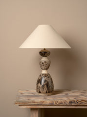 A table lamp with a cream fabric drum shade and a sculptural double-bulb base made of polished stone in mottled gray, brown, and cream, displayed on a rustic wooden table against a solid beige wall.
