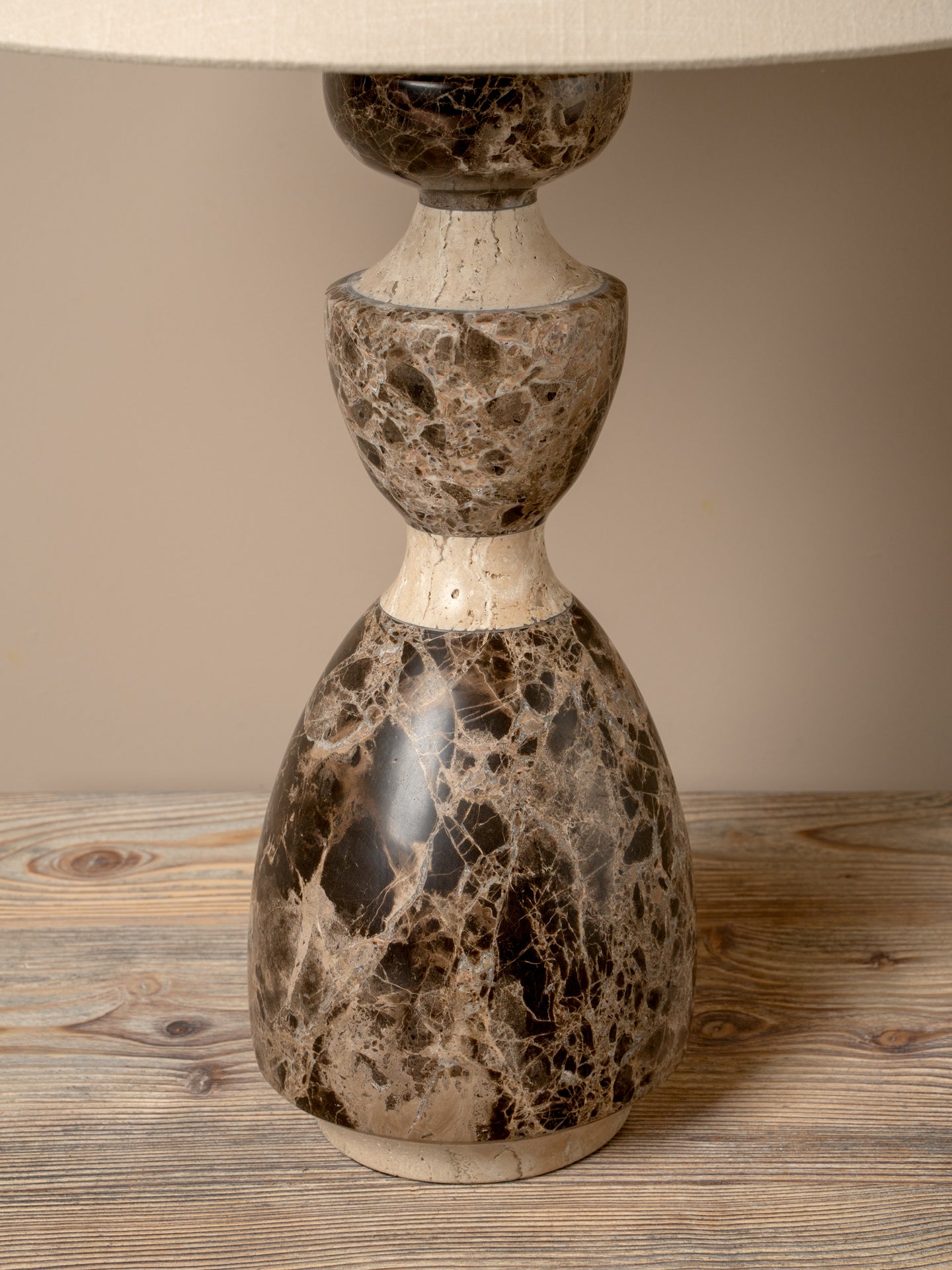 A decorative table lamp with a sculptural, stacked base made of polished stone in a mottled brown and beige marble pattern, topped with a cream fabric drum shade, set on a rustic wooden surface.