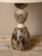 A decorative table lamp with a sculptural, stacked base made of polished stone in a mottled brown and beige marble pattern, topped with a cream fabric drum shade, set on a rustic wooden surface.