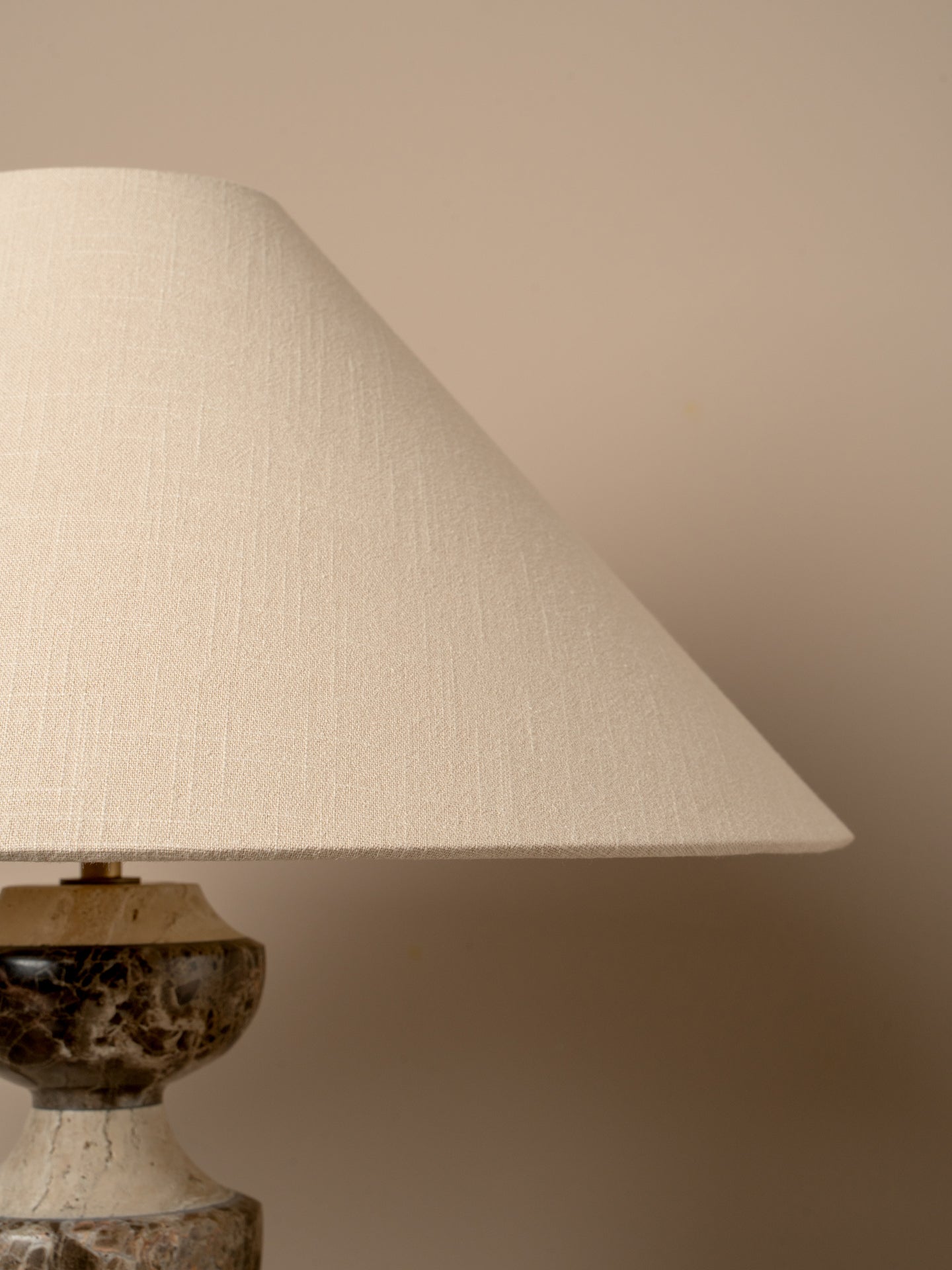 A close-up of a table lamp with a large tapered off-white linen shade and a dark marbled stone base against a warm beige background.