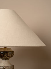 A close-up of a table lamp with a large tapered off-white linen shade and a dark marbled stone base against a warm beige background.
