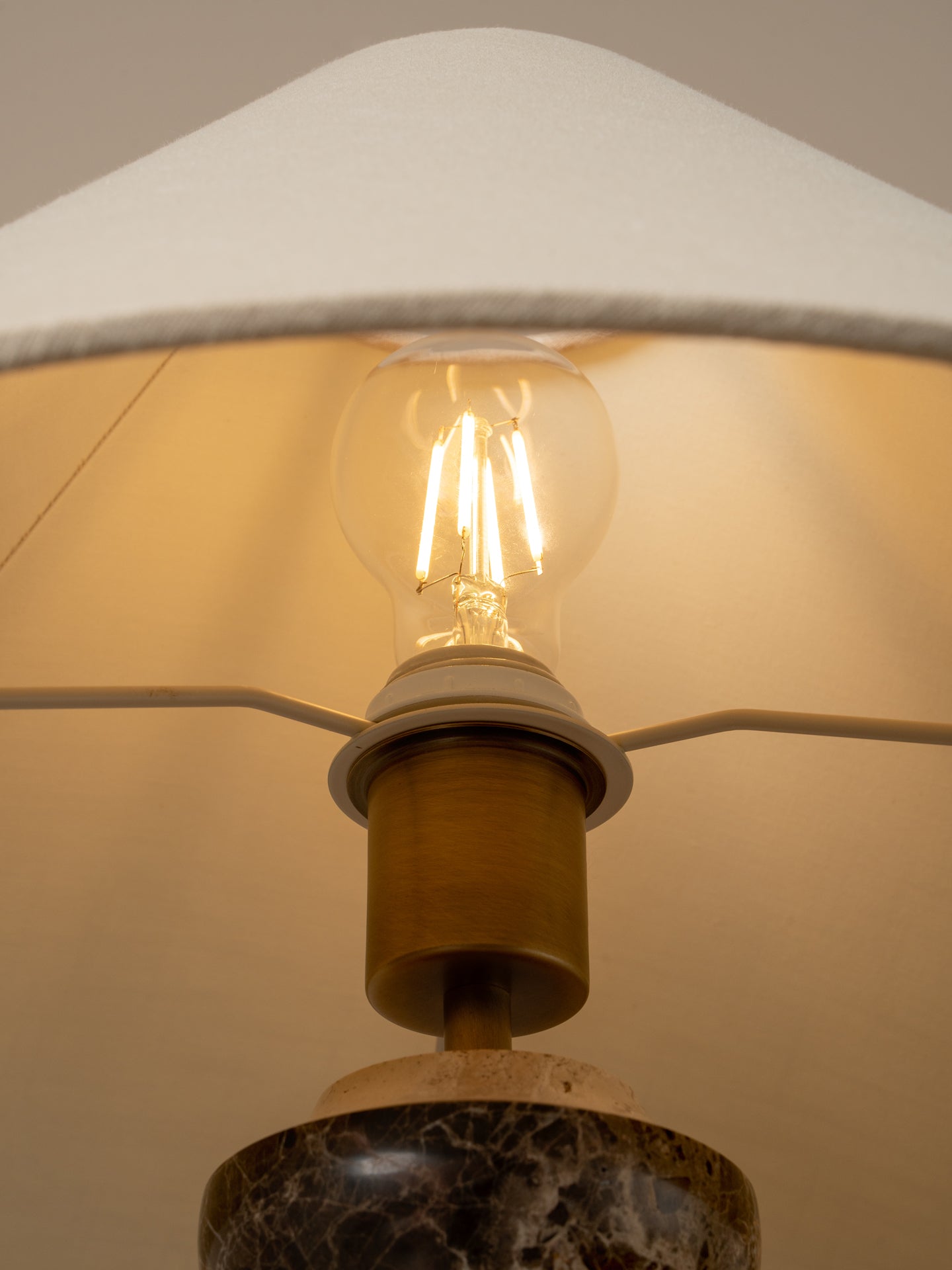 Close-up of a table lamp with a warm glowing Edison bulb, a brass-colored metal neck, and a dark marble base under a white fabric shade.