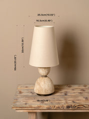 A table lamp with a tapered beige fabric shade and a textured stone-like base sits on a rustic wooden table against a beige wall, with measurement lines and labels showing its dimensions.