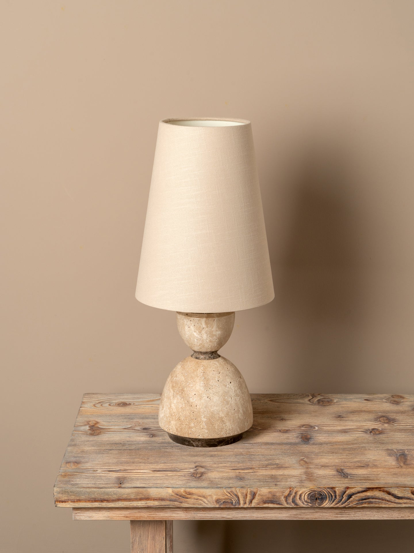 A table lamp with a tapered off-white fabric shade and a textured, rounded beige stone-like base, displayed on a rustic wooden table against a solid beige wall.