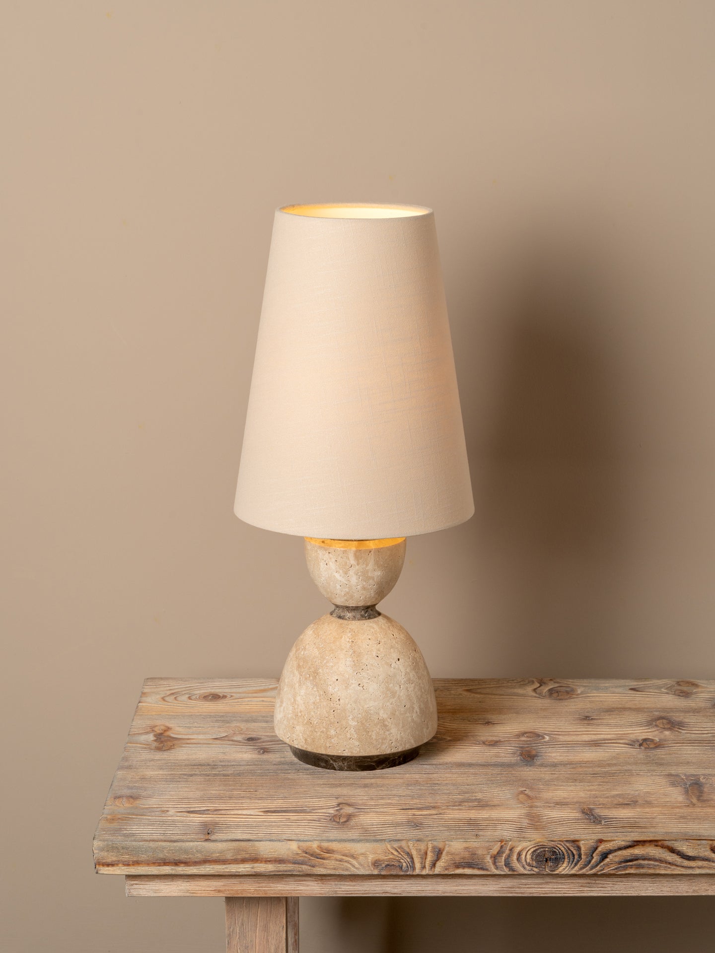 A table lamp with a tapered beige fabric shade and a textured, stone-like beige and cream base, shown lit on a rustic wooden table against a solid beige wall.