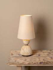 A table lamp with a tapered beige fabric shade and a textured, stone-like beige and cream base, shown lit on a rustic wooden table against a solid beige wall.