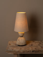 A rustic table lamp with a textured beige fabric drum shade and a carved stone-like base, illuminated with a warm yellow glow, set on a weathered wooden table against a solid taupe background.