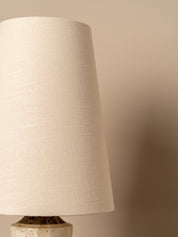 A close-up of a table lamp featuring a large, tapered off-white linen shade and a textured stone base against a warm beige background.