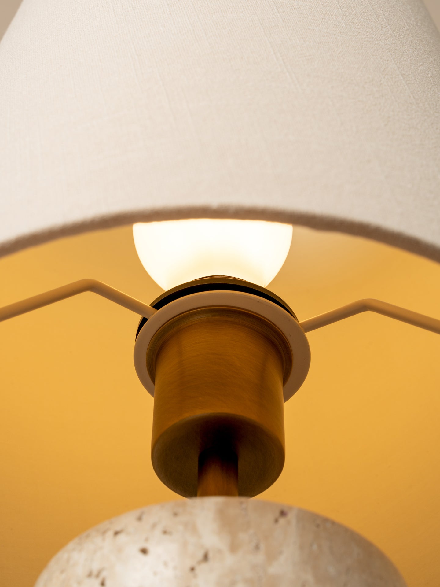 Close-up of a modern pendant light with a textured off-white fabric drum shade, a warm brass cylindrical socket, and a white speckled stone or concrete base, illuminated from within.