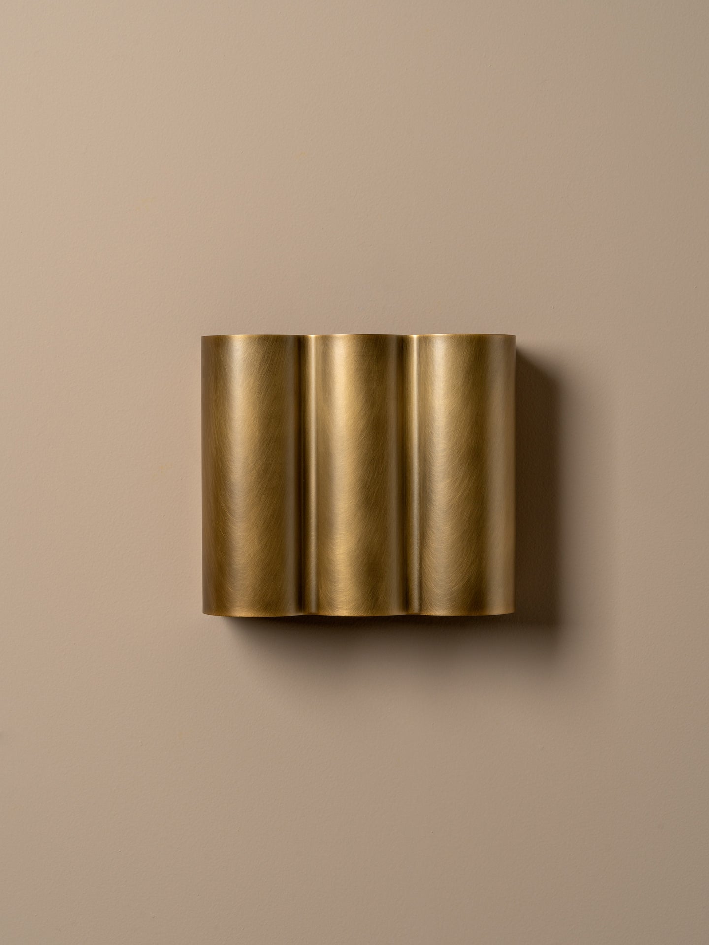 A modern three-cylinder wall sconce in brushed gold metal, mounted on a solid beige wall.