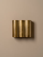 A modern three-cylinder wall sconce in brushed gold metal, mounted on a solid beige wall.