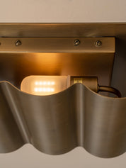 Close-up of a modern wall sconce with a wavy brushed metal shade and a warm glowing light bulb, mounted against a neutral wall.